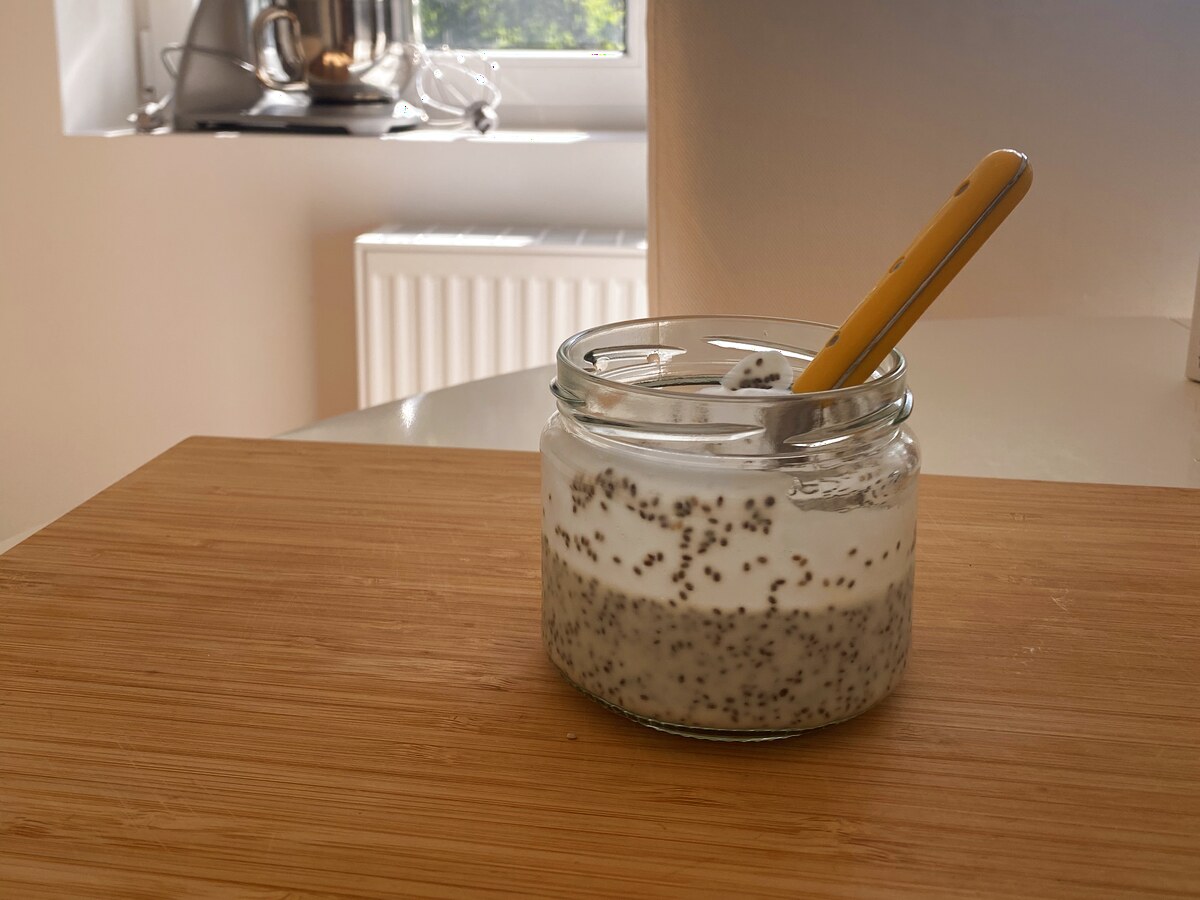 Glazen potje chiapudding met een lepel erin, op een houten tafel bij het raam