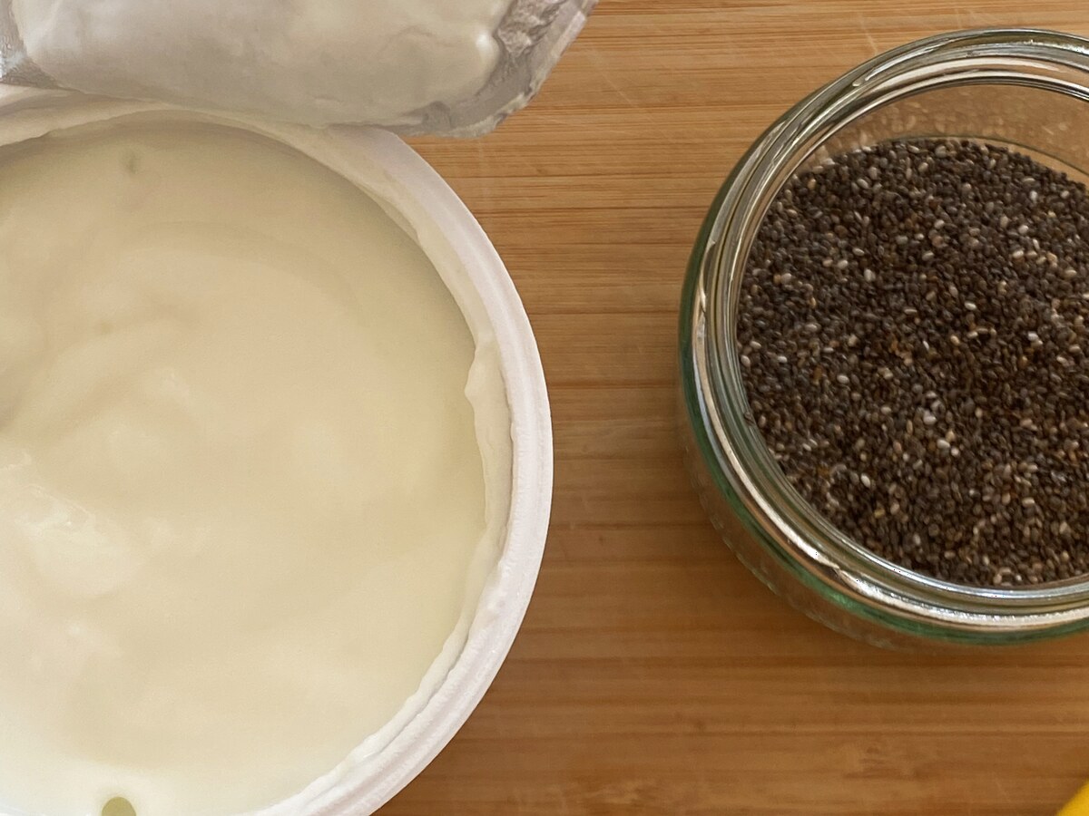 Een open pot yoghurt naast een glazen potje met chiazaad op een houten plank